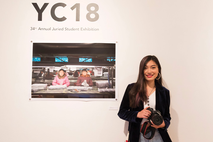 a woman standing next to a picture hanging off the wall with a camera in her hands.